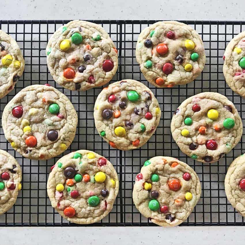 A grid cooling rack filled with freshly baked cookies topped with colorful chocolate candies in red, yellow, green, orange, and brown. The cookies are evenly spaced on the rack.