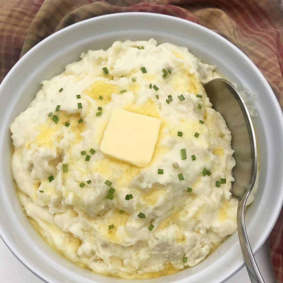 a white serving dish filled with roasted garlic mashed potatoes topped with butter and chopped chives