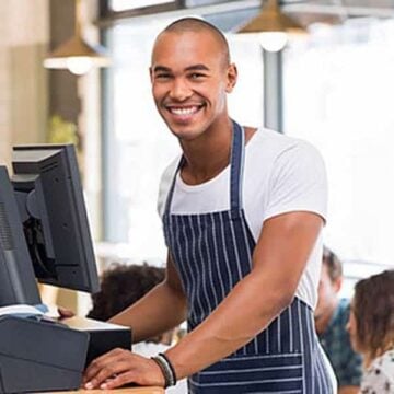 A smiling person wearing a striped apron stands at a register in a café. Theyre looking at the camera, with soft lighting and people in the background.
