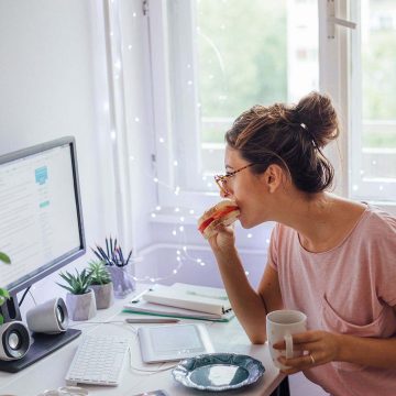 A woman looking at a frugal living blog on a computer monitor while eating a sandwich and holding a coffee cup.