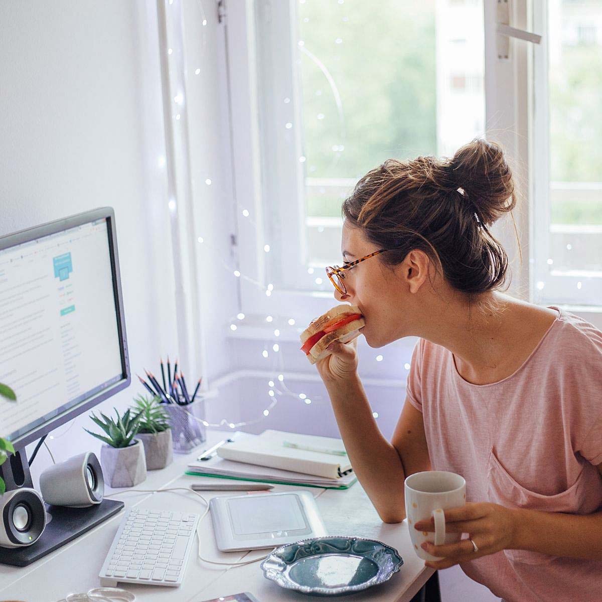 A woman eating a sandwich and reading a blog post on a desktop computer.