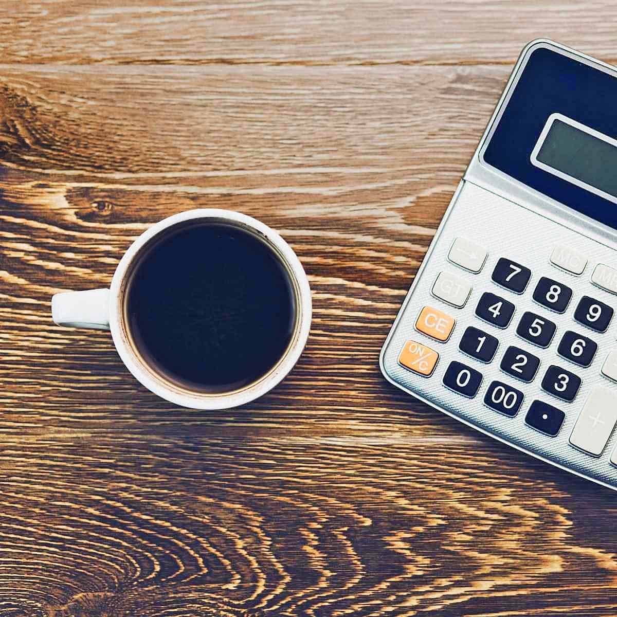 A cup of coffee in a white mug and a calculator on a wood desk.
